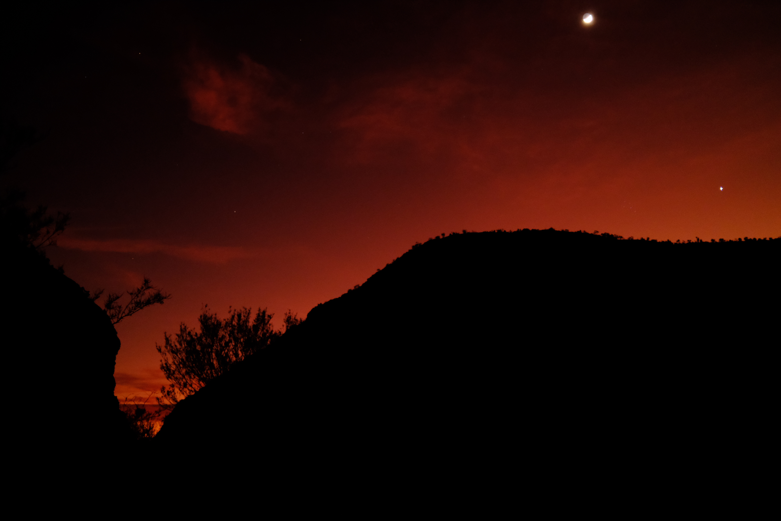 A photograph of the moon and one planet in the sky at dawn over cliffs in silhouette.
